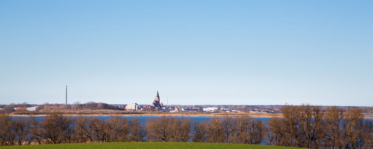 Im Vordergrund ein grünes Feld. In der Mitte ein großes Gewässer, der Bodden. Im Hintergund die Stadt Barth unter bleuem Himmel